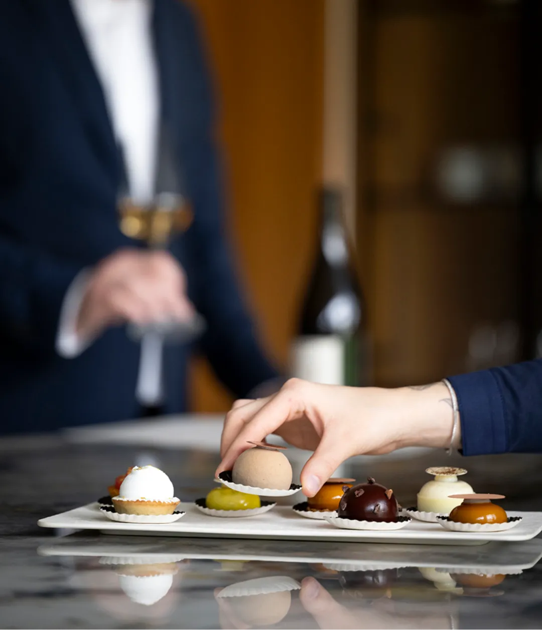 Verfijnde mini patisserie bij uw wijnbeleving. Een hand pakt een ronde mini patisserie van een schaal met diverse desserts tijdens een wijnbeleving.