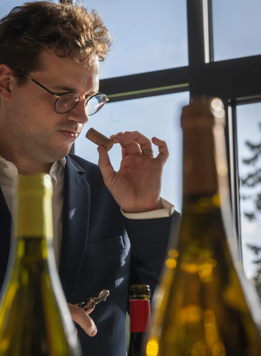 Een sommelier met bril en blauw pak ruikt aandachtig aan een wijnkurk met 'PIQUERAS' erop, omringd door flessen.